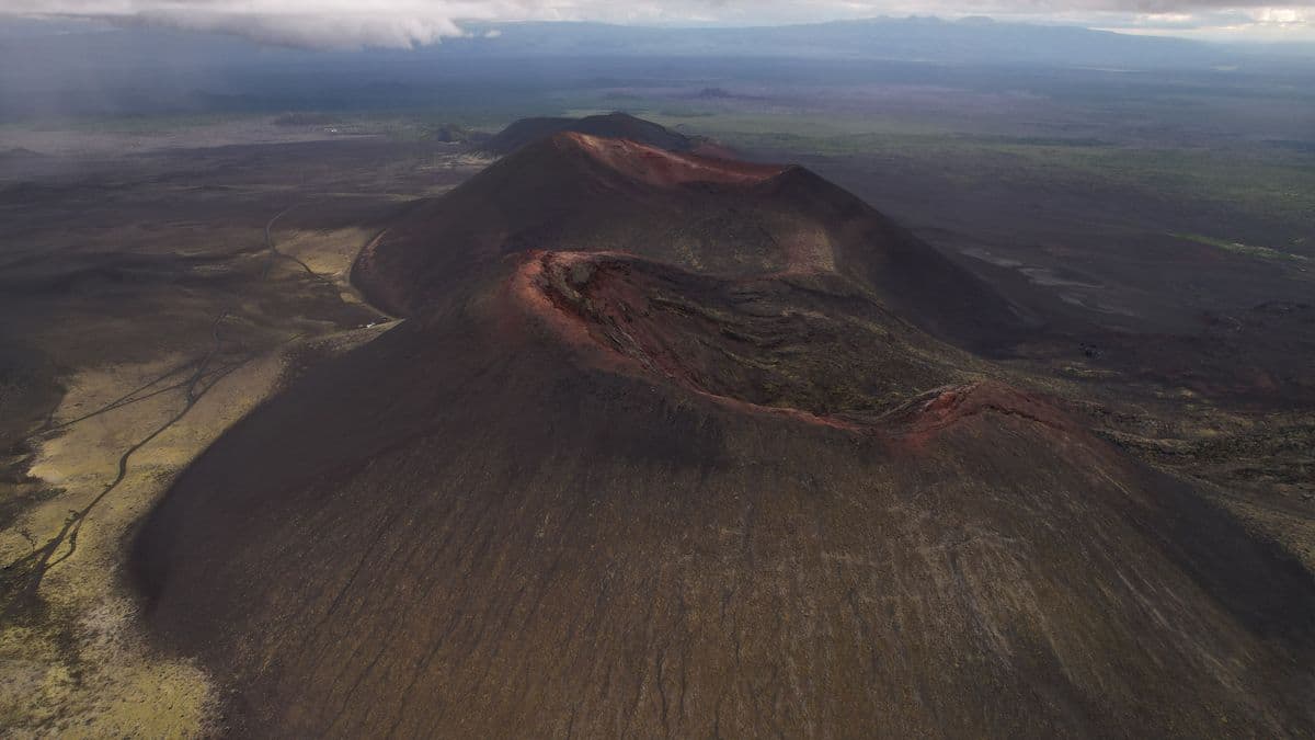 Aerial view of a dark, conical volcano with a reddish crater rim in a barren landscape.