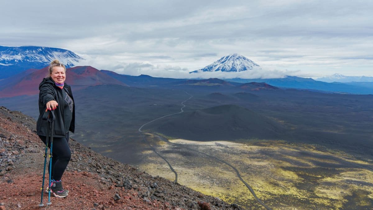 Woman smiling with trekking poles on a volcanic slope overlooking mountains.