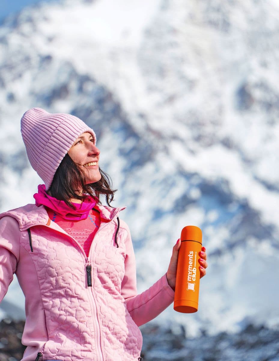Hiker in pink jacket and hat holding a thermos, gazing at a snowy peak.