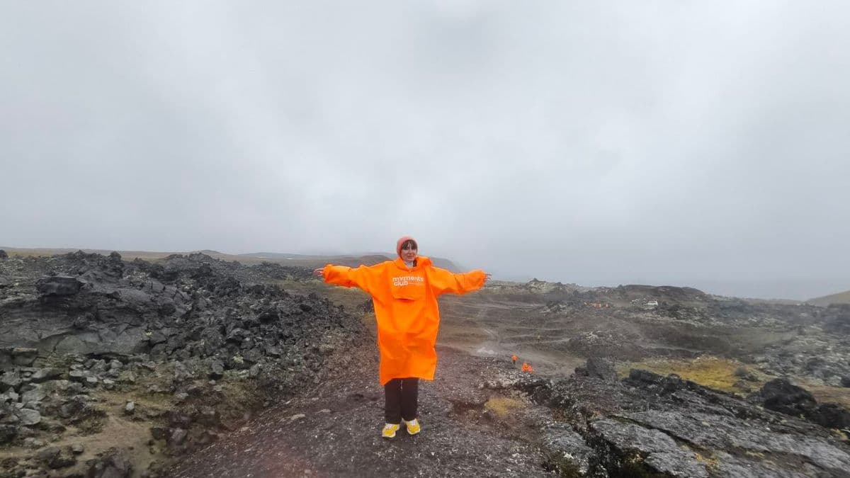 Person in orange poncho stands with arms outstretched on a dark, rocky volcanic landscape.
