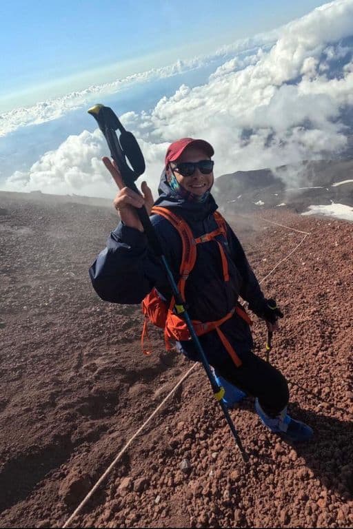 Smiling climber with trekking pole on a high mountain peak above clouds.