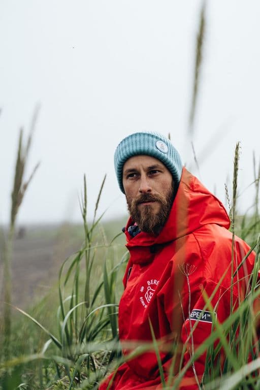 Bearded man in red jacket and blue beanie sits in tall green grass, looking right.