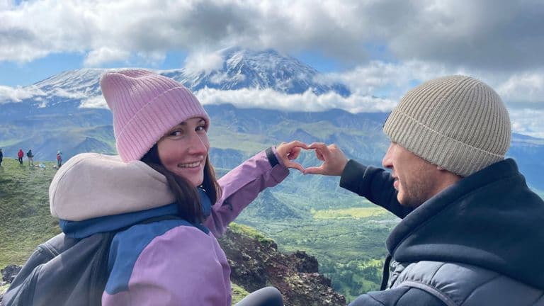 A couple forms a heart with their hands, framed against a majestic snow-capped mountain.