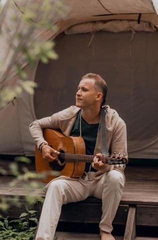 Man playing acoustic guitar outdoors, sitting on a deck beside a tent.
