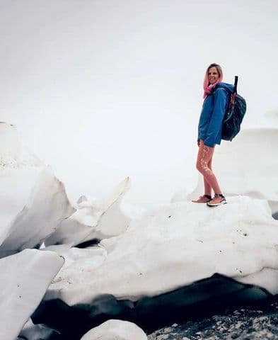 Person with pink hair, blue jacket, and backpack stands on a large ice chunk.
