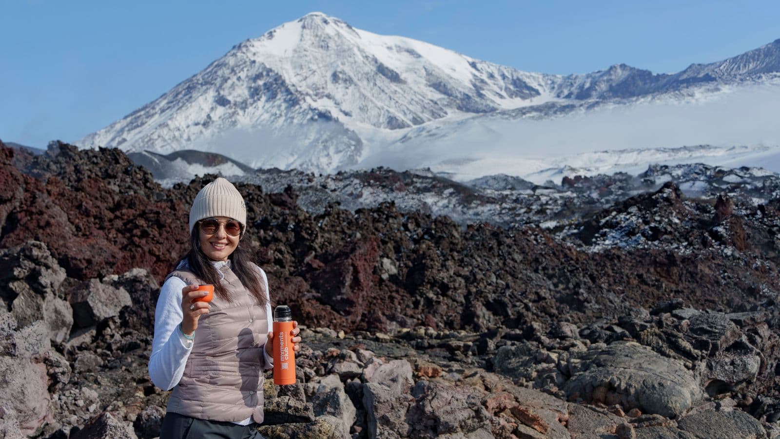Woman with fruit and bottle in volcanic landscape, huge snow-capped mountain in background.