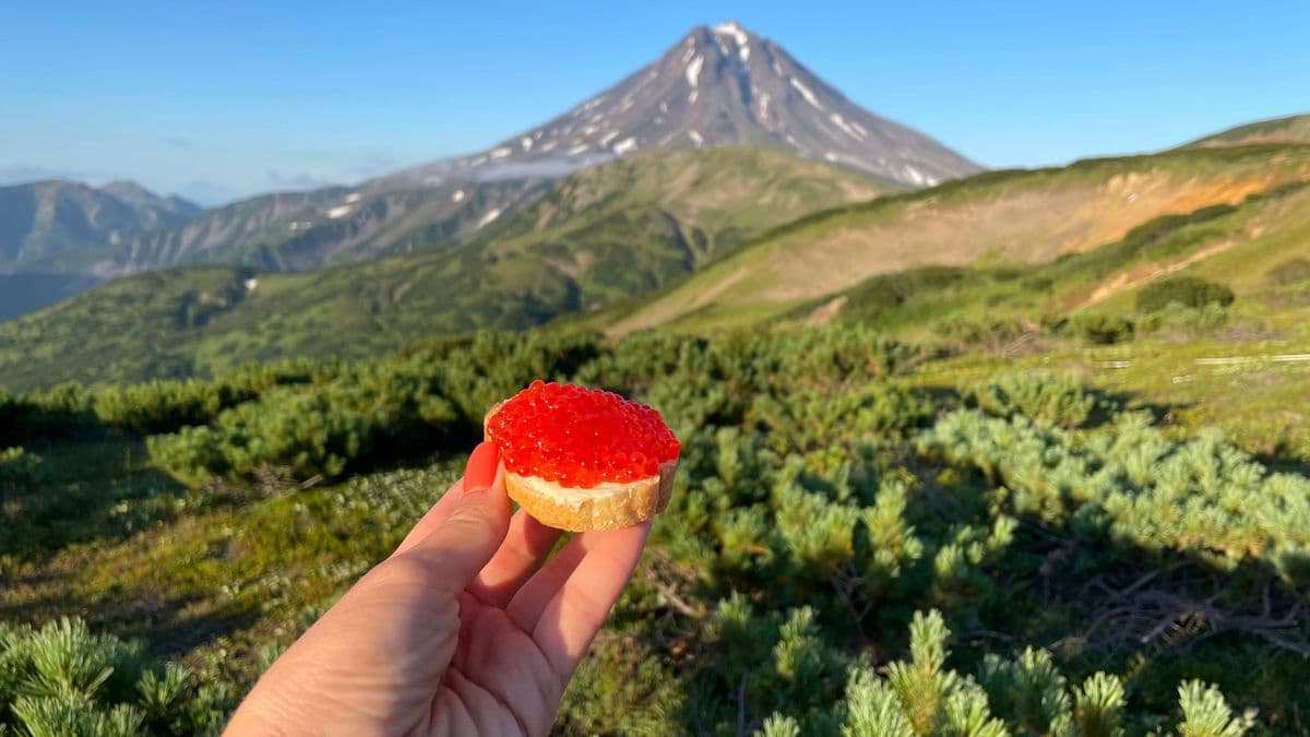 Hand holding bread with red caviar against a stunning volcanic mountain landscape.
