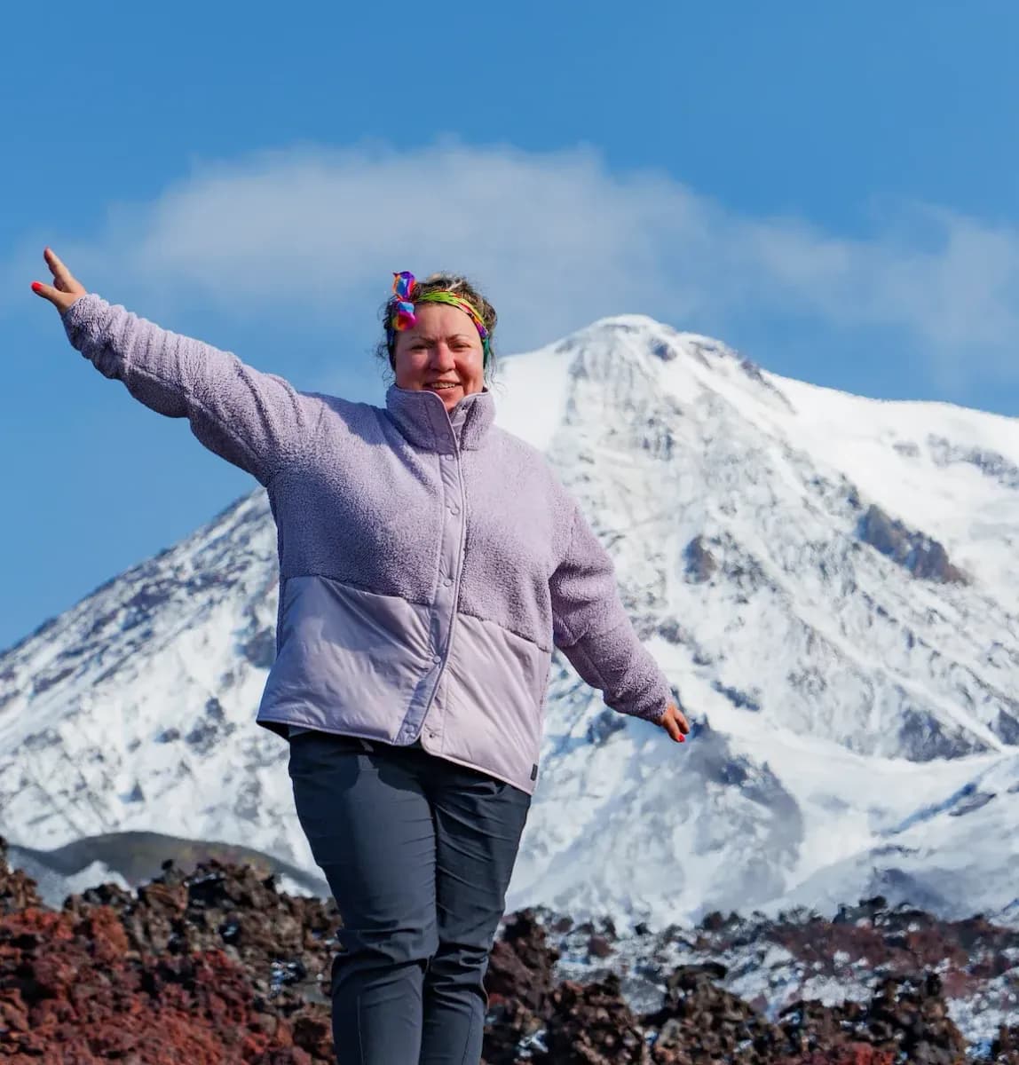 Smiling woman in a purple jacket with outstretched arms before a snow-capped mountain.