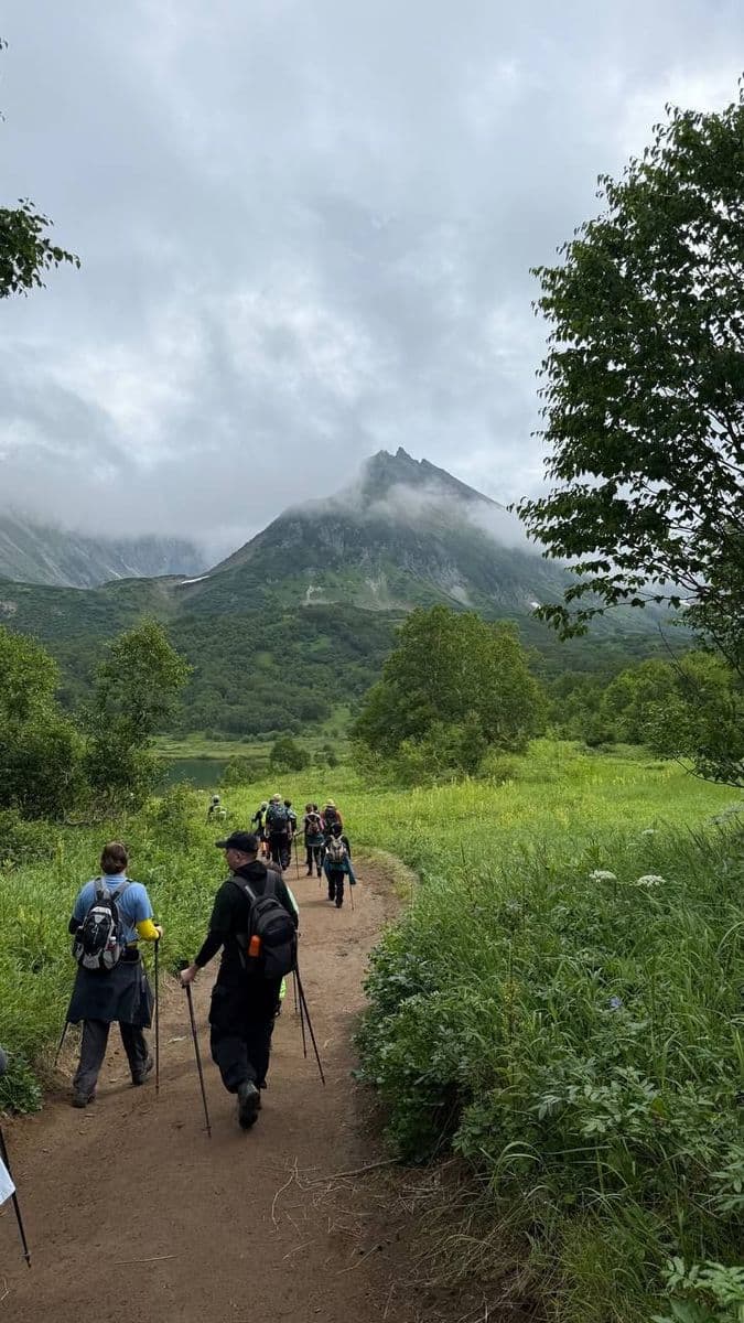 A group hikes a dirt path through green fields, towards misty mountains.
