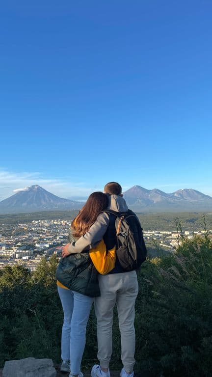 Couple embracing on a hilltop, gazing at a town and distant mountains under blue sky.