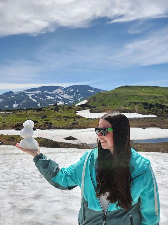 Woman in sunglasses holds a small snowman in a snowy mountain landscape.