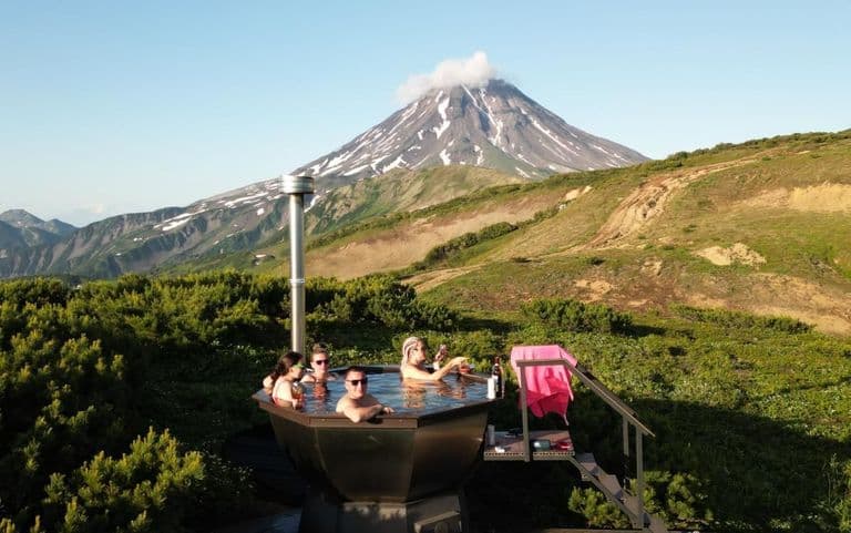 Four people relax in an outdoor hot tub overlooking a majestic volcano under clear skies.