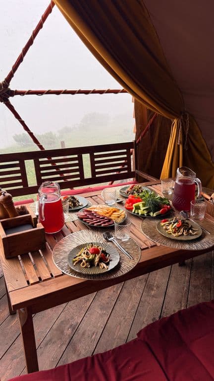 Rustic table in glamping tent with food and drinks, misty mountain view.