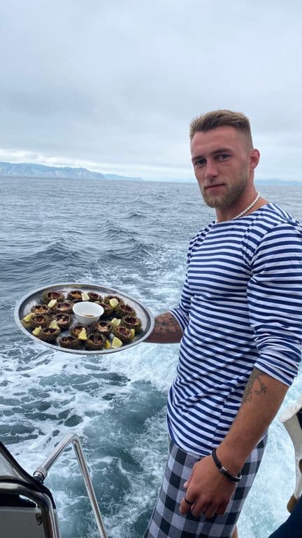 Man in striped shirt on a boat, holding a tray of appetizers with the sea behind him.
