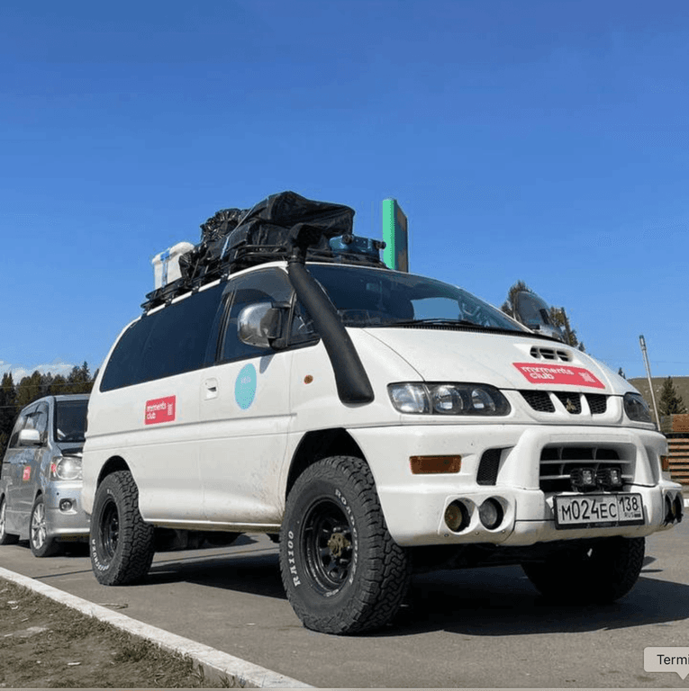 White Mitsubishi Delica minivan, highly modified for off-road adventures, featuring large tires and a roof rack.