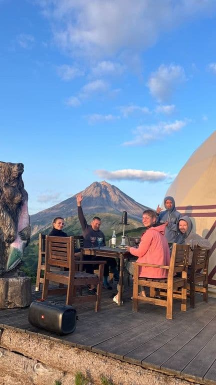 Five friends sit outdoors at a wooden table with a large mountain in the background.