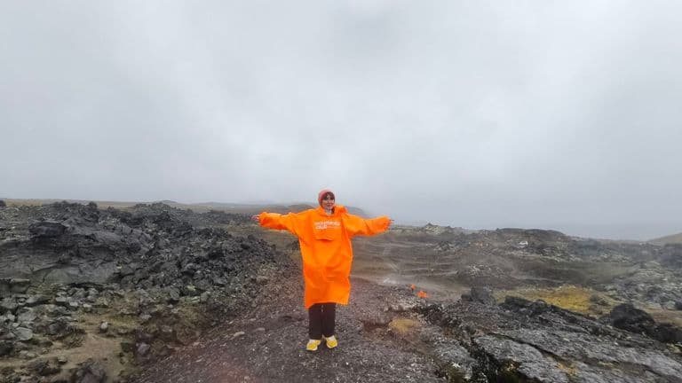 Person in orange poncho stands with arms outstretched on a dark, rocky volcanic landscape.