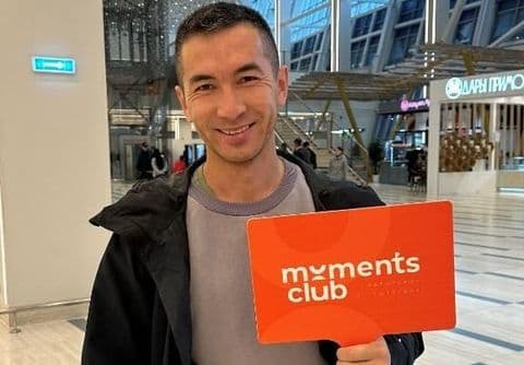Man smiles holding an orange 'moments club' sign inside a modern building.
