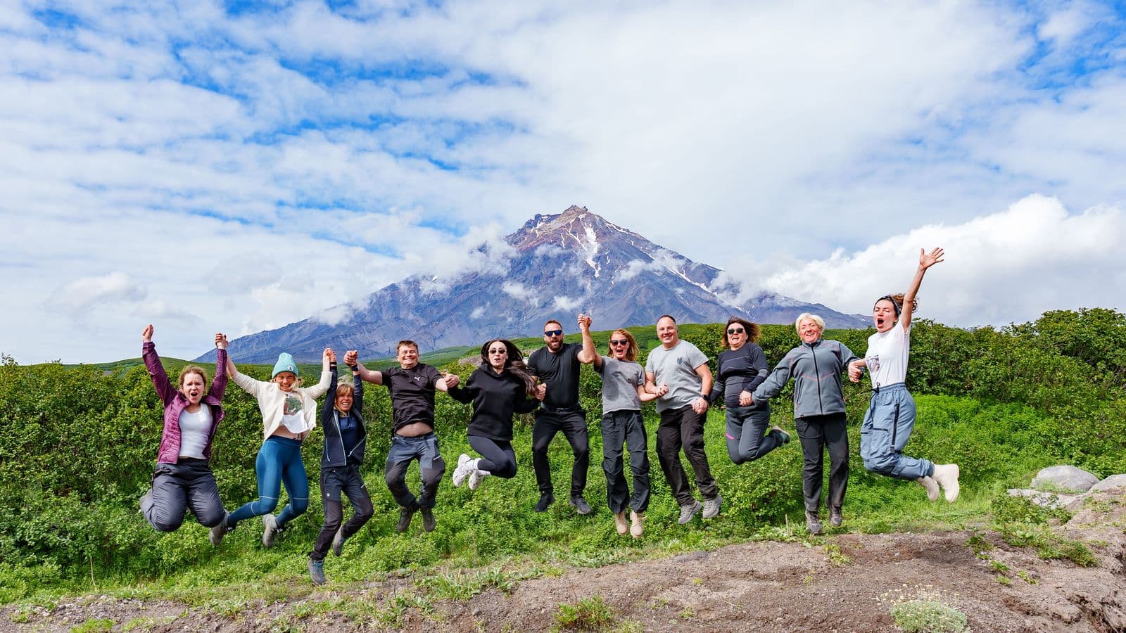 Joyful group jumps high on a green hill with a cloud-covered volcanic mountain in background.